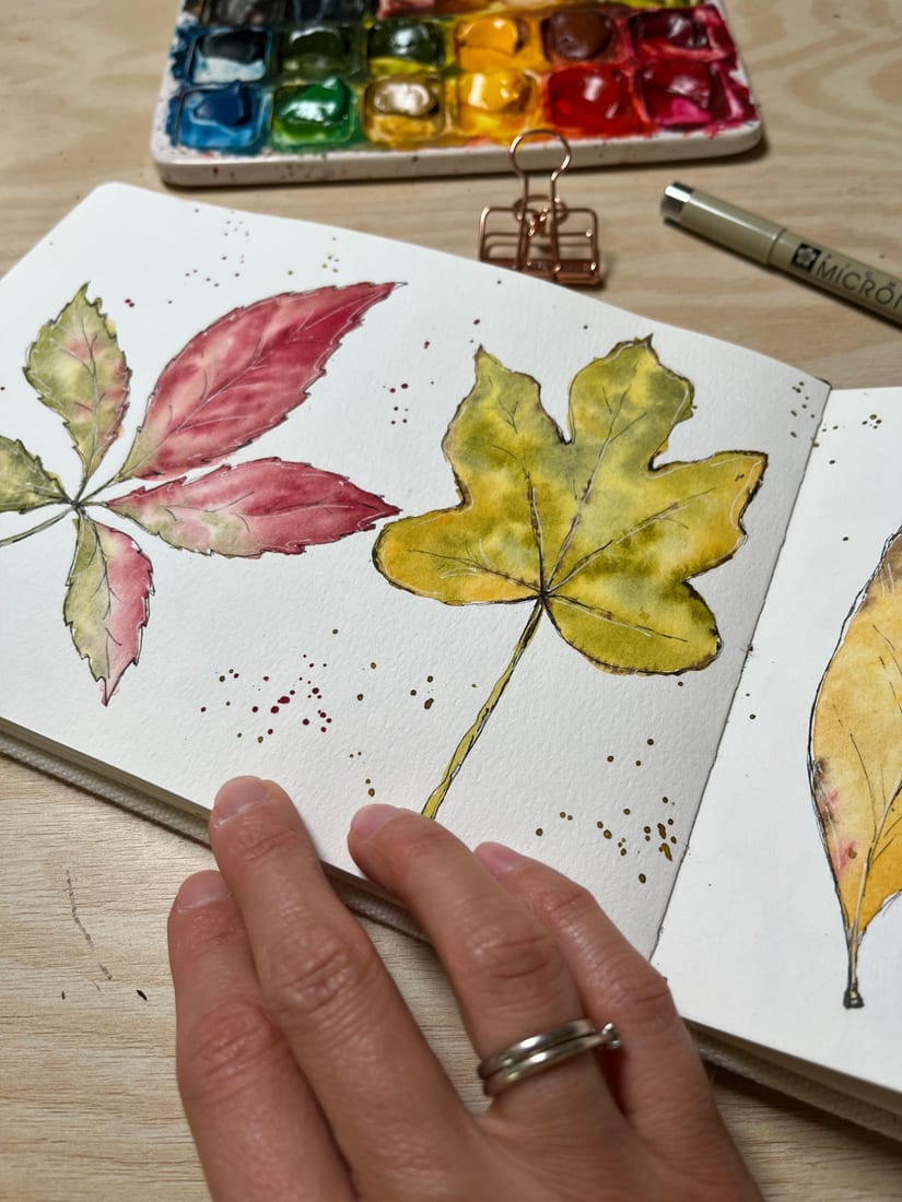 A sketchbook spread of painted autumn leaves in red, green and yellow, with a watercolor palette and fineliner pen
