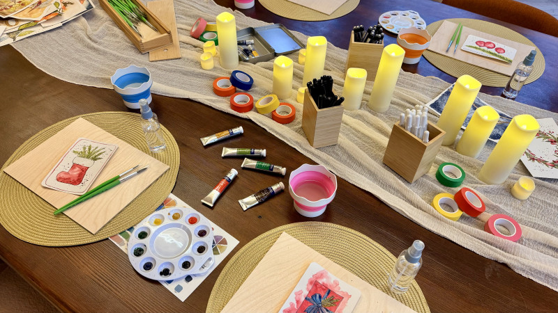 A workshop table set with glowing candles, paint tubes, palettes and a Christmas card at every place
