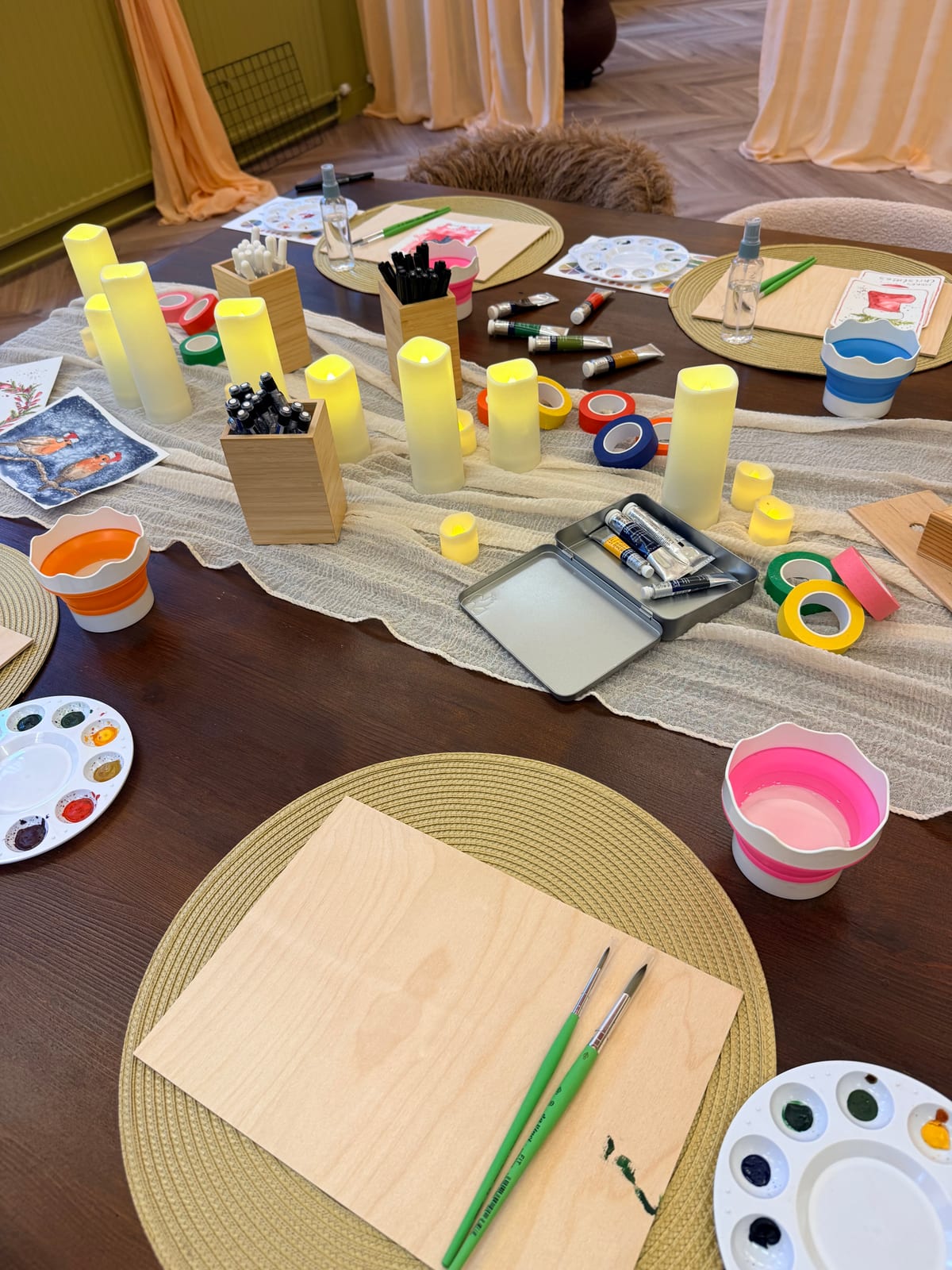 A long workshop table with glowing candles, paint tubes and a palette at each place