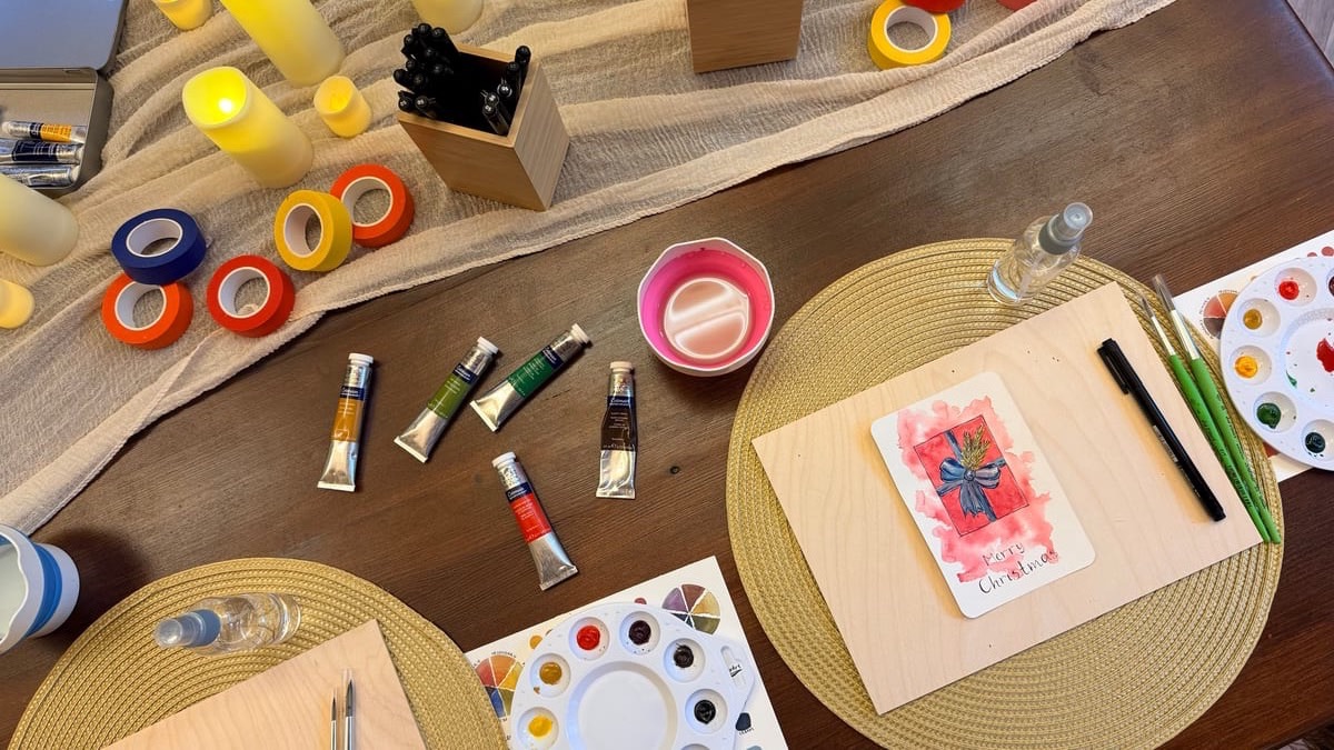 Looking down on paint tubes, washi tape and a painted Christmas card on a wooden board